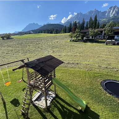 Ein Spielplatz mit einer Rutsche und Schaukeln steht in einer grünen Wiese. Im Hintergrund sind Berge und Häuser zu sehen.