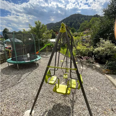 A playground with a yellow swing and a trampoline in the background. The surroundings are green and mountainous.