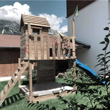 A wooden playground with a slide and a viewing platform is located in the garden. In the background, mountains and the bright facade of a house can be seen.