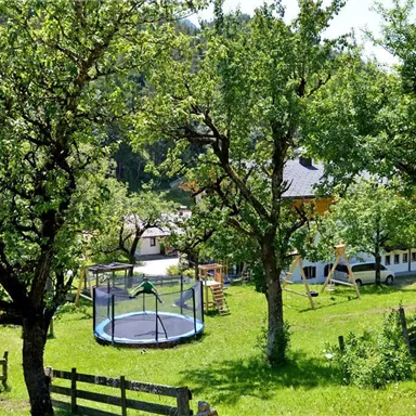 A beautiful garden with many trees and a large trampoline. In the background, a playground and a building can be seen.