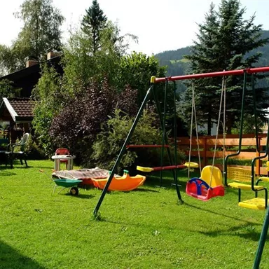 Ein schöner Garten mit einer Schaukel und einem Spielbereich. Im Hintergrund sind Bäume und ein Haus zu sehen, umgeben von Bergen.