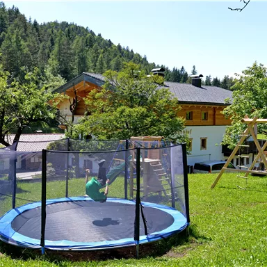 A trampoline on a green meadow, surrounded by trees and a playground. In the background, there is a large, bright house.