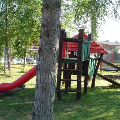 A playground with a slide and a wooden play tower. Green grass and trees surround the play equipment.