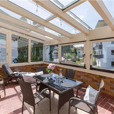 A sunlit conservatory with a table and chairs. In the background, there are green plants and modern residential buildings.