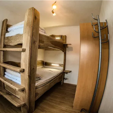 Ein einfaches Zimmer mit einem Holzstockbett und einem Kleiderschrank. Es hat warmes Licht und eine gemütliche Atmosphäre.