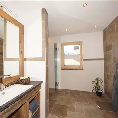 A modern bathroom with a large mirror and elegant stone tiles. The light comes in through a window, and there is a decorative plant arrangement.
