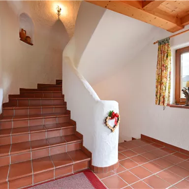 Ein gemütlicher Flur mit einer Treppe, die nach oben führt. Das Licht scheint durch ein Fenster und eine farbige Gardine verleiht dem Raum einen freundlichen Ausdruck.