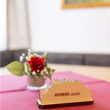 A table with a red tablecloth, on which there is a small flower vase with a rose and a decorative wooden figure with the inscription "GOING strengthens!" In the background, a blurred image can be seen.
