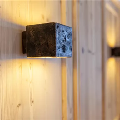 A modern wall lamp made of metal emits a warm light. The background consists of light wood, creating a cozy atmosphere.