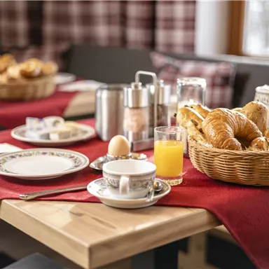 A beautifully set breakfast table with a basket full of fresh croissants. In addition, cups, glasses, and a red tablecloth for a cozy atmosphere.