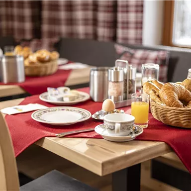 A cozy breakfast table with fresh rolls, croissants, and a glass of orange juice. The tables are decorated with red tablecloths, and there is a selection of dishes and drinks.