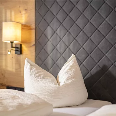 A cozy bedroom with a padded headboard and bright pillows. Next to the bed, there is a wall lamp.