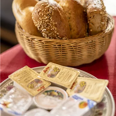 A basket with various rolls is on a table. Next to it, several small portions of butter and jam are arranged on a plate.