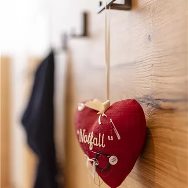 A red fabric heart with the inscription "Emergency" hangs on a hook on a wooden wall. Next to it, dark garments are hanging.