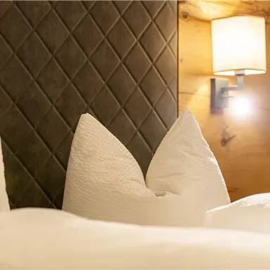 A cozy bed with large, white pillows and a modern bedside lamp. The wall is made of wood and features an elegant, diagonal pattern.