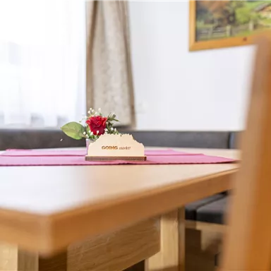 A wooden chair and a table with a red tablecloth. On the table, there is a small flower decoration with a card.
