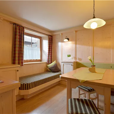 A cozy living room with wood paneling, a table, and seating. A window provides daylight and a clock is mounted on the wall.