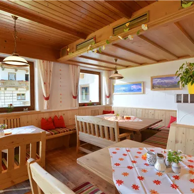 A cozy restaurant with wooden furniture and warm lighting. The windows offer a view of the surroundings, while the tablecloths are decorated with floral patterns.