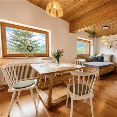 A cozy dining area with a wooden table and white chairs. Large windows offer a beautiful view of nature.