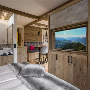 Ein modernes Zimmer mit Holzmöbeln und einem gemütlichen Bett. Im Hintergrund ist ein Fernseher mit einer Landschaft zu sehen.