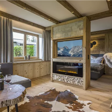 Ein gemütlicher Wohnraum mit Holzdetails und einem modernen Kamin. Es gibt eine Sitzecke mit Wein und einem Blick auf die Berge.