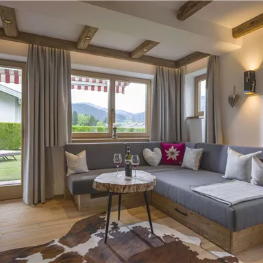 Ein gemütlicher Wohnbereich mit einer großen Sofaecke und einem Kaffeetisch. Große Fenster bieten einen Blick auf den Garten und die Berge.