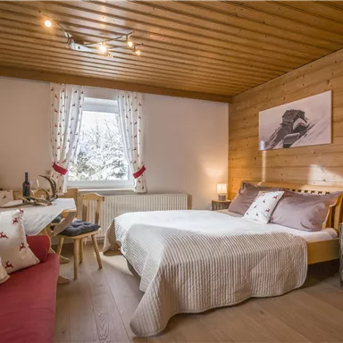 A cozy room with a wooden ceiling and wooden walls. There is a double bed, a small seating area, and large windows that let in daylight.