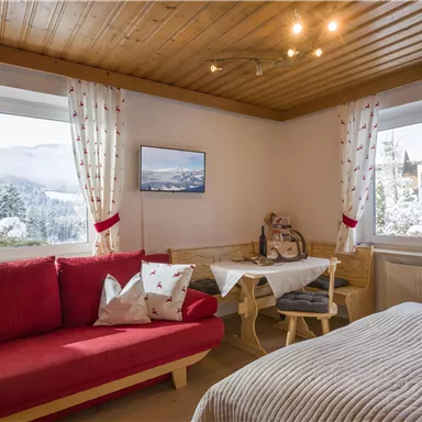 A cozy room with a red sofa and a bed. Through the window, one looks out onto a wintry landscape.