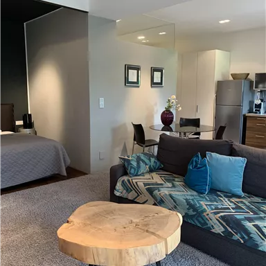 Ein modernes Wohnzimmer mit einem grauen Sofa und einem runden Tisch aus Holz. Im Hintergrund ist ein Schlafzimmer mit einem bequemen Bett zu sehen.