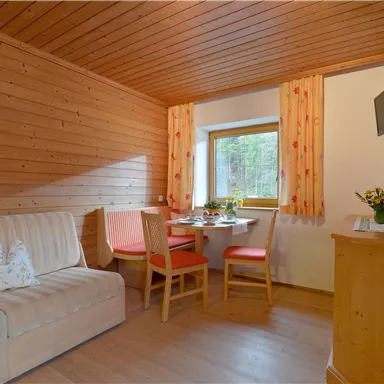 A cozy room with wooden walls and a sofa. There is a dining area with a wooden table and chairs, as well as a TV on the wall.