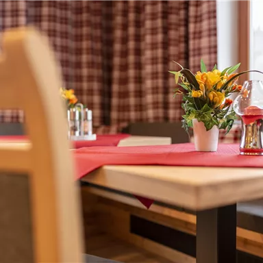 An inviting table with a red tablecloth, a floral arrangement, and a glass vase. In the background, cozy windows and a checked pattern are visible.