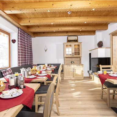 A cozy breakfast room with wooden ceilings and large windows. Tables are set with fresh rolls, drinks, and dishes.