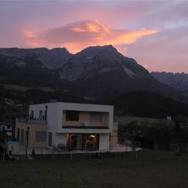 Ein modern aussehendes Haus steht vor einer beeindruckenden Berglandschaft. Der Himmel ist in sanften Farben bei Sonnenuntergang gefärbt.