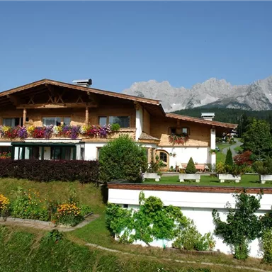 Ein charmantes alpenländisches Haus mit Blumenbalkonen, umgeben von grünen Wiesen. Im Hintergrund sind majestätische Berge zu sehen.