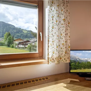 Ein heller Raum mit einem Fenster, das eine grüne Landschaft und Berge zeigt. Neben dem Fenster steht ein Fernseher, der ebenfalls eine Aussicht auf die Natur zeigt.