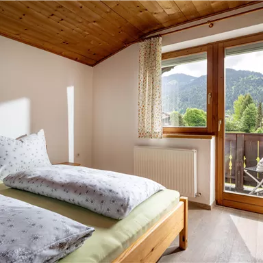 Ein helles Schlafzimmer mit zwei Betten und einer großen Fensterfront. Im Hintergrund sind Berge und ein Balkon mit Stühlen zu sehen.