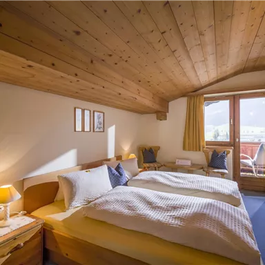 Ein gemütliches Schlafzimmer mit zwei Betten und einer Holzdecke. Große Fenster sorgen für viel Tageslicht und bieten einen Blick nach draußen.