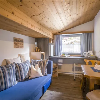 Ein gemütlicher Wohnraum mit blauen Sofakissen und einem Holzregal. Am Fenster steht ein Tisch mit Stühlen und einer kleinen Blumenvase.