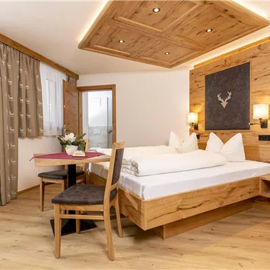 A cozy hotel room with wooden decor and a large window. The room features a double bed, a table, and chairs.