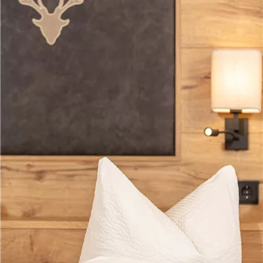 A cozy bedroom with white pillows and a wooden wall. A deer antler and a wall lamp are hanging on the wall.