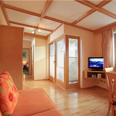 A bright living room with a cozy sofa and a television. The room features wooden details and large windows that let in daylight.