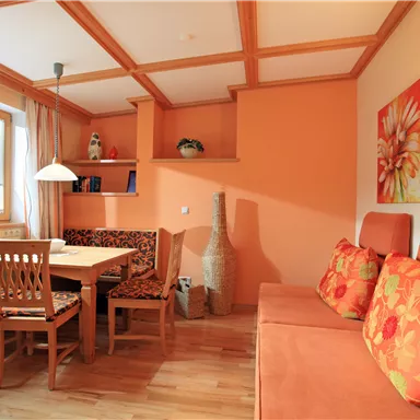 A cozy living space with a dining table and colorful chairs. The walls are painted in warm shades of orange and decorated with floral pictures.