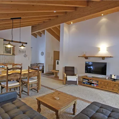 Ein modernes Wohnzimmer mit Holzdecke und einem großen Esstisch. Die gemütliche Einrichtung besteht aus einer Sofagruppe und einem Fernseher.