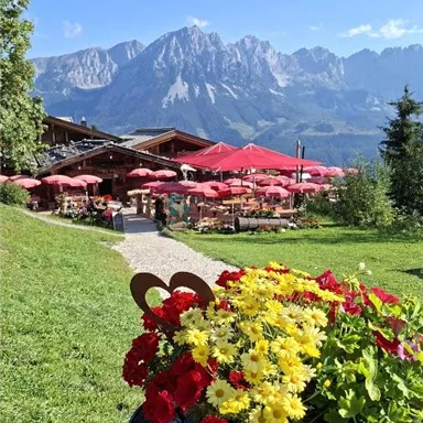 Ein malerisches Restaurant mit roten Sonnenschirmen und blühenden Blumen im Vordergrund. Im Hintergrund erheben sich majestätische Berge unter einem klaren blauen Himmel.