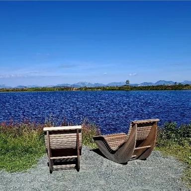 Zwei gemütliche Liegestühle stehen am Ufer eines ruhigen Gewässers. Es ist ein sonniger Tag mit klarem Himmel und grüner Umgebung.