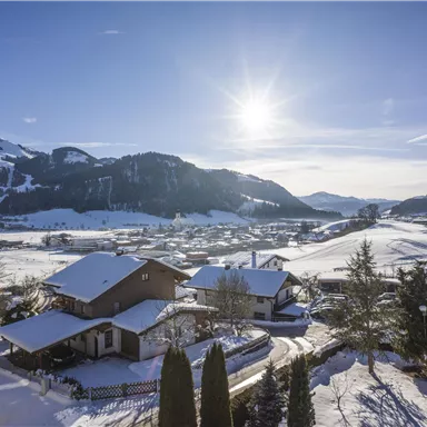 Eine winterliche Landschaft mit schneebedeckten Bergen und strahlendem Sonnenlicht. Kleine Häuser liegen in einem idyllischen, verschneiten Tal.