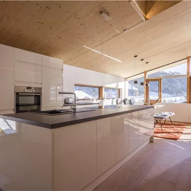 Eine moderne Küche mit weißen Möbeln und großen Fenstern, die viel Tageslicht hereinlassen. Im Hintergrund ist eine beeindruckende Berglandschaft zu sehen.