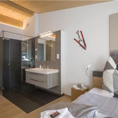 Ein modernes Schlafzimmer mit Holzakzenten und einem angrenzenden Badezimmer. Die Einrichtung ist schlicht und elegant, mit einem großen Spiegel und einem gemütlichen Bett.