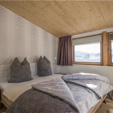 Ein gemütliches Schlafzimmer mit einem Doppelbett und zwei großen Kissen. Durch die Fenster hat man einen Blick auf die Berge und die umgebende Landschaft.