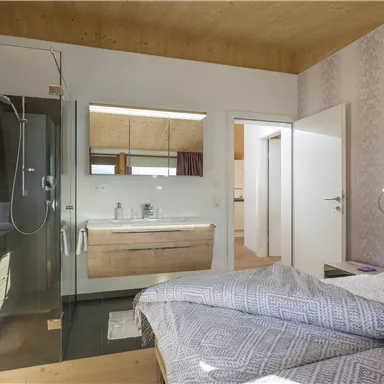 Ein modernes Schlafzimmer mit einem Badezimmerbereich. Die Dusche ist durch eine Glastür getrennt und der Raum wirkt hell und einladend.